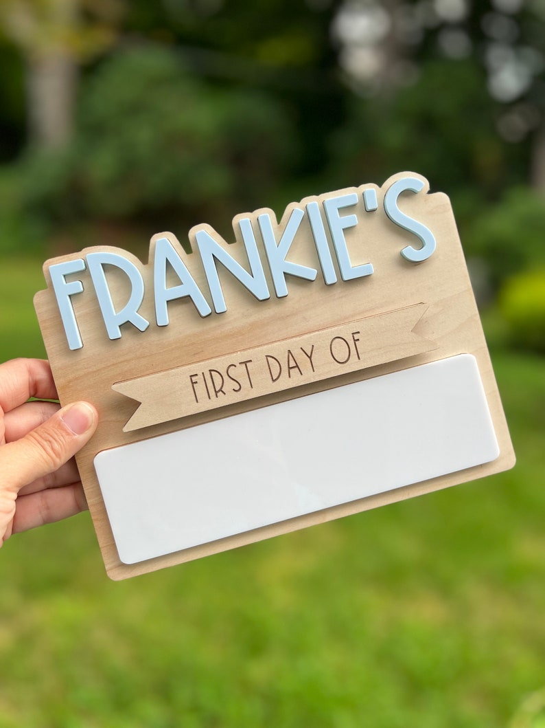 May include: A wooden sign with the name "FRANKIE'S" in blue letters and a banner that says "FIRST DAY OF" with a blank space for writing. The sign is held in a hand.