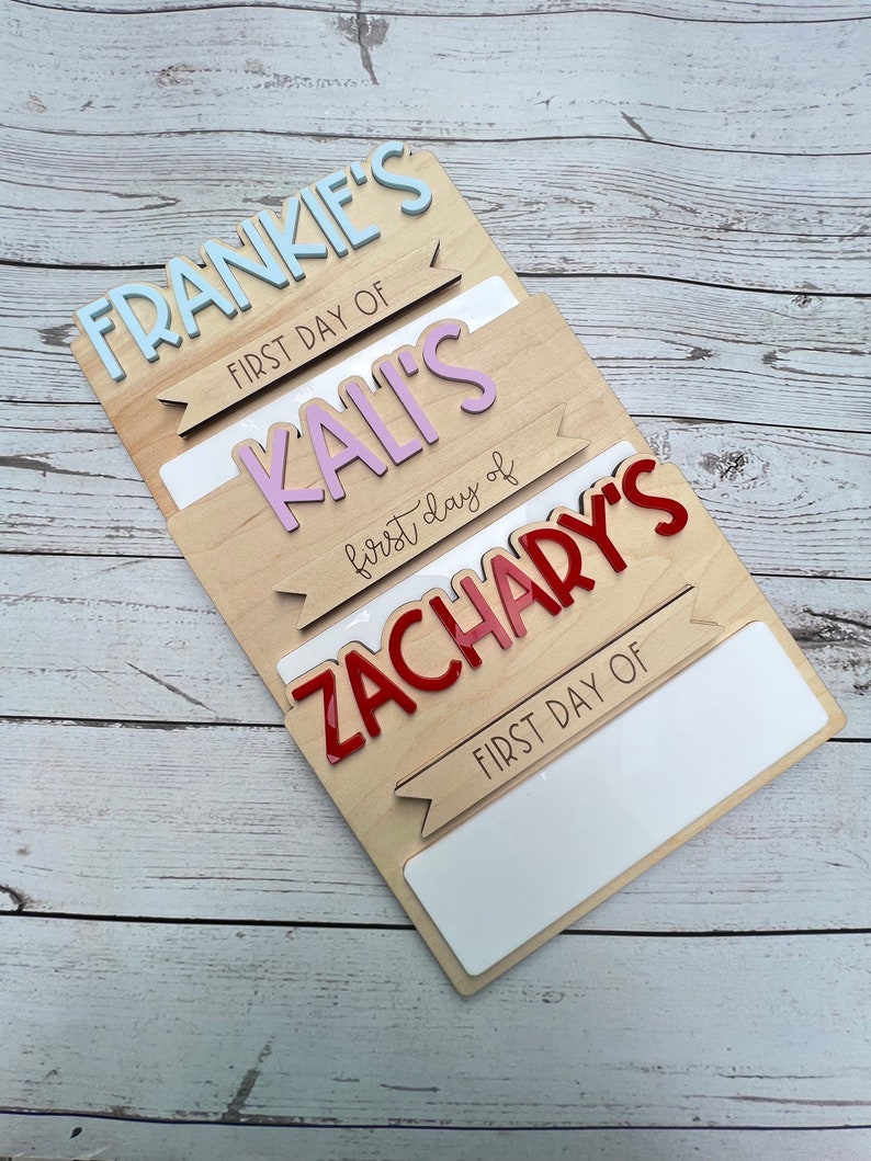 May include: Three wooden signs with the names Frankie, Kali, and Zachary, each with a banner that says "First Day Of". The signs are layered on top of each other, with the top sign being Frankie's and the bottom sign being Zachary's.