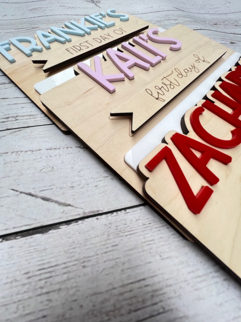 May include: Three wooden signs with the names Frankie, Kail, and Zachariah, each with the text "first day of" in cursive. The signs are stacked on top of each other, with Frankie's sign on top and Zachariah's sign on the bottom. The names are in different colors: Frankie's name is light blue, Kail's name is pink, and Zachariah's name is red.