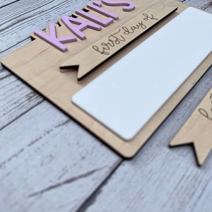 May include: A wooden sign with the name "KAILI'S" in pink letters and the text "first day of" written in black. The sign has a blank white space for writing in the center.
