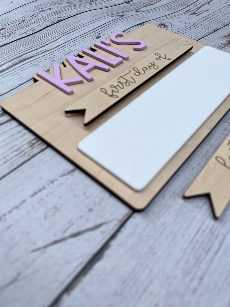 May include: A wooden sign with the name "KAILI'S" in pink letters and the text "first day of" written in black. The sign has a blank white space for writing in the center.