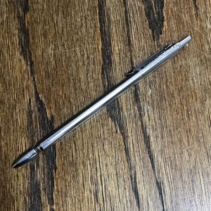 May include: A slim, metallic silver mechanical pencil featuring a hexagonal barrel design, a pointed tip, and a pocket clip attached to the upper section. The writing instrument has a sleek, polished finish and a simple, functional construction.