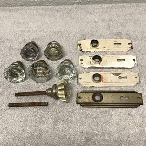 May include: A collection of six clear glass door knobs with faceted edges, accompanied by four rectangular metal door plates. Three plates feature a distressed white finish, while one plate has a dark, weathered metallic surface. Two metal spindles are included.