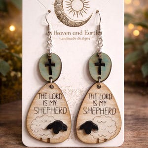 May include: Wooden dangle earrings with a religious theme. Each earring features a cross on a blue-green oval and a teardrop shape with a sheep illustration and the text "THE LORD IS MY SHEPHERD". The earrings are suspended from silver-colored hooks.
