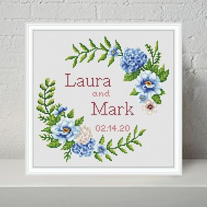 May include: Cross stitch pattern featuring a floral wreath with blue and white flowers, green leaves, and the text "Laura and Mark 02.14.20".