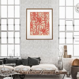 May include: A framed print of a red and white botanical illustration of palm leaves. The print is hanging on a gray wall above a wooden bench with black cushions.