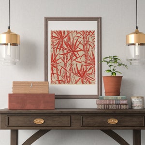 May include: A framed print of a red and white tropical leaf design hangs above a wooden console table with two drawers. Two gold pendant lights hang above the table. A small potted plant sits on the table next to a stack of books.