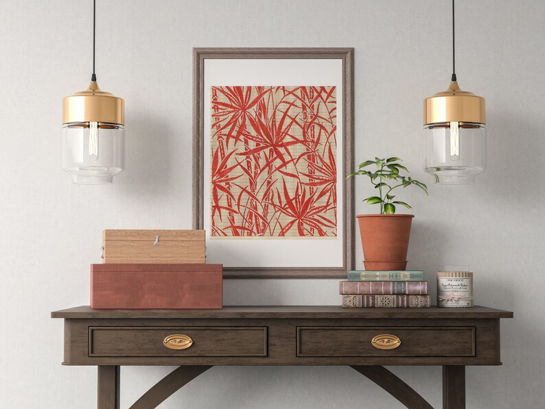 May include: A framed print of a red and white tropical leaf design hangs above a wooden console table with two drawers. Two gold pendant lights hang above the table. A small potted plant sits on the table next to a stack of books.