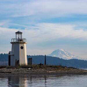 May include: A white lighthouse with a black roof and a circular window stands on a small island. A snow-capped mountain is visible in the background under a cloudy blue sky. The lighthouse is surrounded by water.