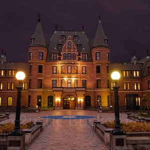 May include: A nighttime exterior shot of a large, ornate brick building with multiple turrets and a dark roof. The building is illuminated by warm interior lights and street lamps, set against a deep purple sky. A brick walkway leads to the entrance.
