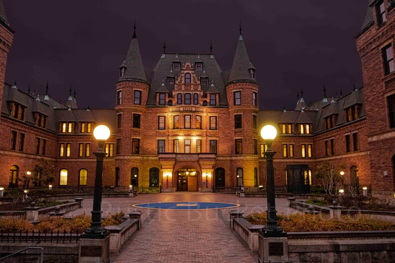 May include: A nighttime exterior shot of a large, ornate brick building with multiple turrets and a dark roof. The building is illuminated by warm interior lights and street lamps, set against a deep purple sky. A brick walkway leads to the entrance.