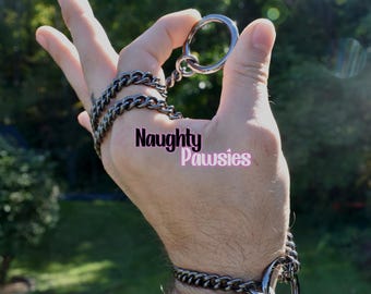 May include: A silver metal ring and chain set. The set includes a large ring held by a hand, connected to a chain that wraps around the wrist. The text "Naughty Pawsies" is displayed in pink.