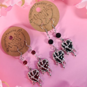 May include: A pair of silver heart-shaped earrings with black and red resin. The hearts are dripping with silver and have a chain design. The earrings are hanging from silver hooks with black and red beads.