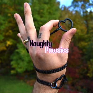 May include: Black heart-shaped restraints with a chain design. The restraints are held in a hand, with the text "Naughty Pawsies" in pink and black. The background is a blurred view of green and red foliage.