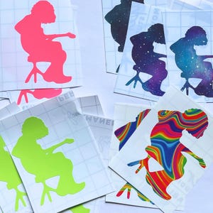 May include: A collection of vinyl stickers featuring a silhouette of a person playing a guitar while seated. The stickers come in various colors and patterns, including neon pink, green, galaxy, and a rainbow swirl design.