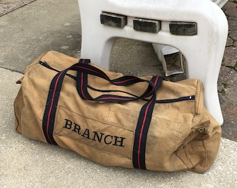 May include: A tan canvas duffel bag with black and red straps. The bag has the word "BRANCH" embroidered on the front.