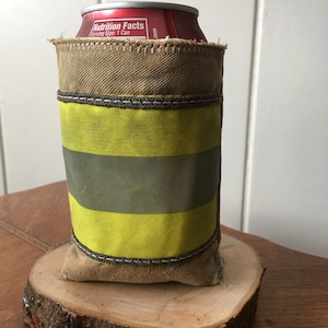 May include: A brown canvas can cooler with a yellow reflective stripe and a red can inside. The cooler is sitting on a wooden slice.