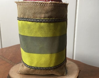 May include: A brown canvas can cooler with a yellow reflective stripe and a red can inside. The cooler is sitting on a wooden slice.