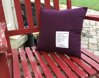 May include: A purple pillow with a white fabric patch sewn on the front. The patch has the text "This is a shirt I used to wear. When you hold it know I am there. Love, Grandma". The pillow is sitting on a red wooden rocking chair.