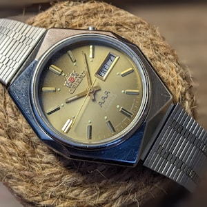 May include: A gold-toned wristwatch with a silver metal band. The watch face has a gold background with black markings and a date window that reads "FREI 7". The watch brand is "Citizen" with the text "AAA" below the brand name.