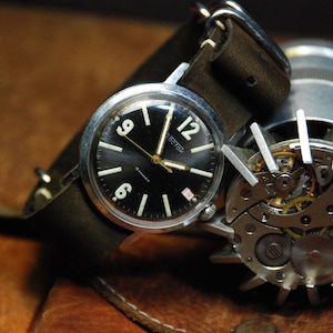 May include: A silver wristwatch with a black face and white markings. The watch has a brown leather band and is sitting on a brown leather surface. The watch face says "Rostok".