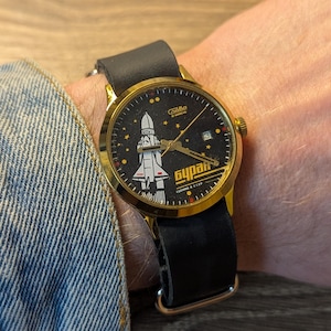 Könnte beinhalten: Eine goldfarbene Armbanduhr mit schwarzem Zifferblatt, das eine weiße und rote Raumfähre und das kyrillische Wort "Бурaн" zeigt. Die Uhr hat ein schwarzes Lederarmband und wird am Handgelenk getragen.