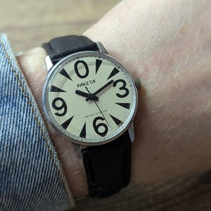 Peut inclure: Une montre-bracelet avec un boîtier rond argenté et un cadran crème présentant de grands chiffres noirs aux positions 0, 3, 6 et 9. La montre possède des aiguilles noires et le texte PAKETA imprimé sur le cadran, avec un bracelet en cuir noir.