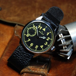 May include: A black leather wristwatch with a black face and yellow numerals. The watch has a small second hand dial and the word "PILOT" printed on the face.