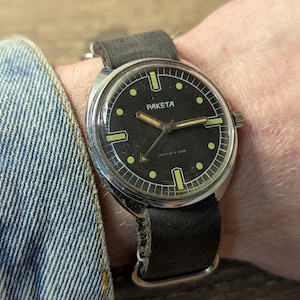 Puede incluir: Un reloj de pulsera de cara negra con una caja plateada y una correa de cuero negra. La esfera del reloj tiene marcas blancas y la palabra "PAKEta" impresa en ella.