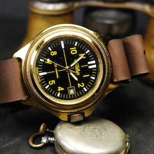 Puede incluir: Un reloj de pulsera dorado con una esfera negra y marcas blancas. El reloj tiene una correa de cuero marrón y una ventana de fecha en la posición de las 4 en punto. La esfera del reloj dice "21 jewels" y "Слава".