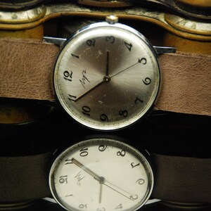 May include: A silver-toned wristwatch with a round face, black Arabic numerals, and a light brown leather strap. The watch dial features the Cyrillic text "Луч" in black script. The watch is positioned above its own reflection on a dark surface.