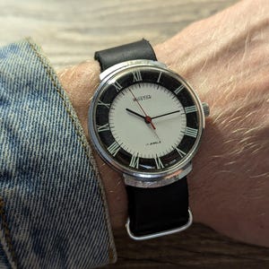 Puede incluir: Un reloj de pulsera plateado con una esfera blanca y marcadores de hora romanos negros. El reloj tiene una correa de cuero negra y se lleva en la muñeca.