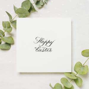 May include: A square, off-white card with the words "Happy Easter" in elegant black script. The card is surrounded by sprigs of green eucalyptus leaves, creating a natural, festive border. The background is a textured, light-colored surface.