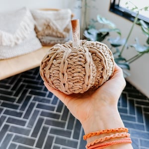 May include: A hand is holding a small, knitted pumpkin with a brown stem. The pumpkin is beige and has a woven texture. The hand is wearing three orange braided bracelets.