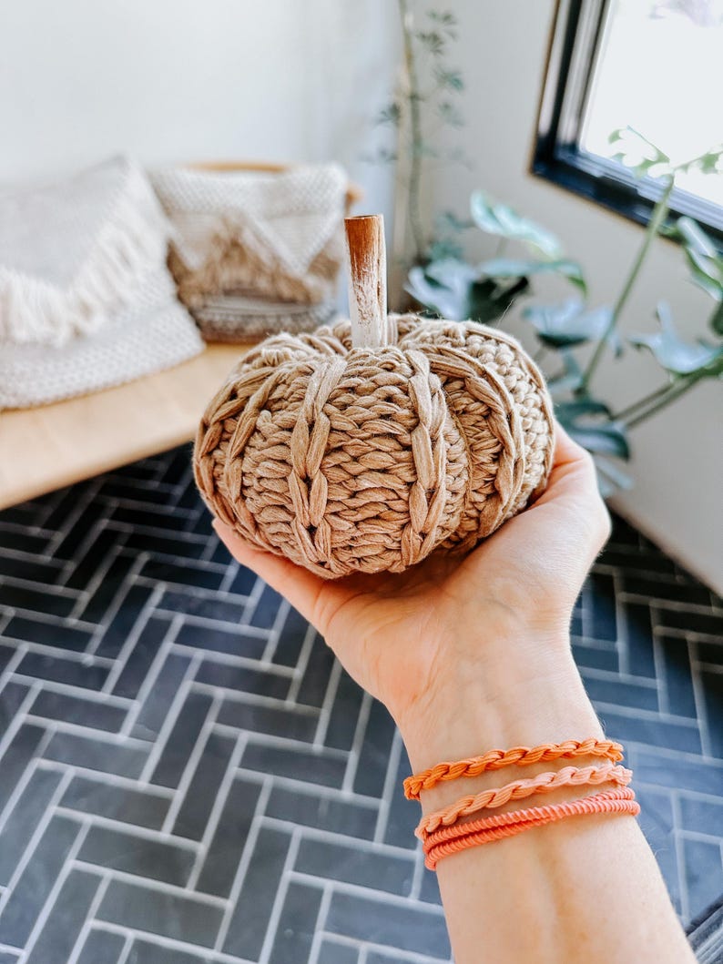 May include: A hand is holding a small, knitted pumpkin with a brown stem. The pumpkin is beige and has a woven texture. The hand is wearing three orange braided bracelets.