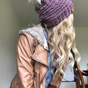 Puede incluir: Un gorro de punto morado con un pomp&oacute;n de piel sint&eacute;tica blanca. El gorro lo lleva una persona con el pelo largo y rubio y una chaqueta de cuero marr&oacute;n.