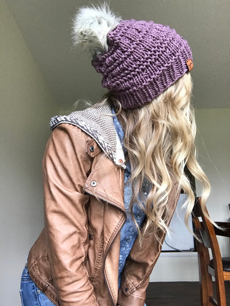 Puede incluir: Un gorro de punto morado con un pomp&oacute;n de piel sint&eacute;tica blanca. El gorro lo lleva una persona con el pelo largo y rubio y una chaqueta de cuero marr&oacute;n.