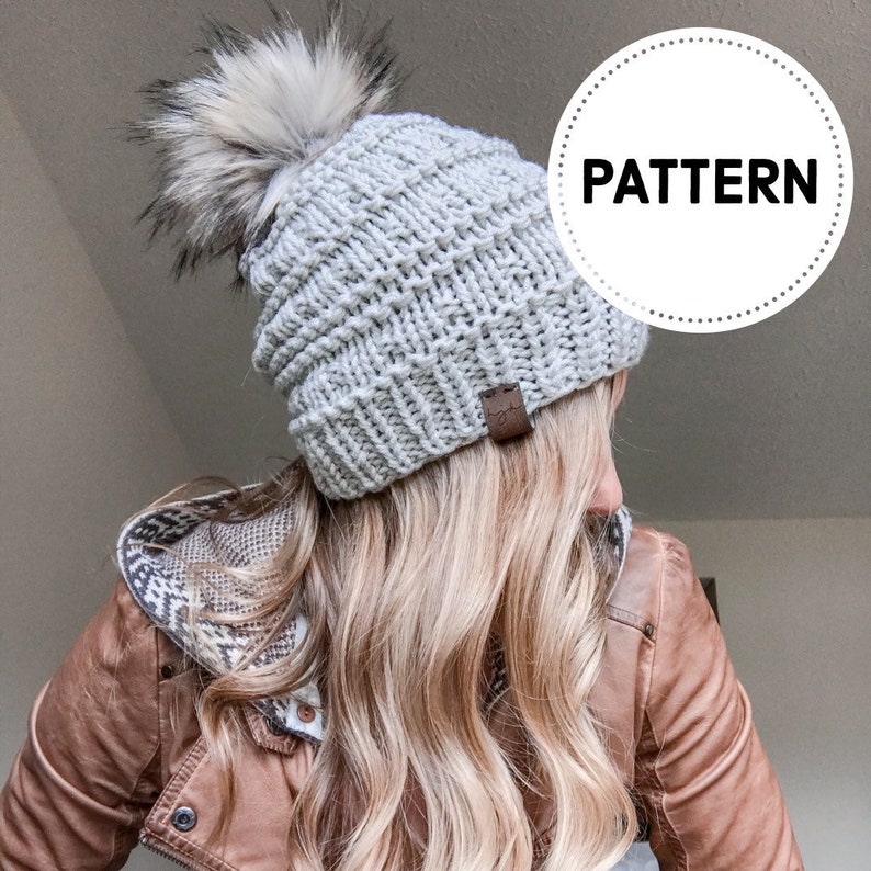 Puede incluir: Un gorro de punto gris con un pomp&oacute;n blanco esponjoso. El gorro tiene una textura acanalada y una etiqueta de cuero marr&oacute;n.