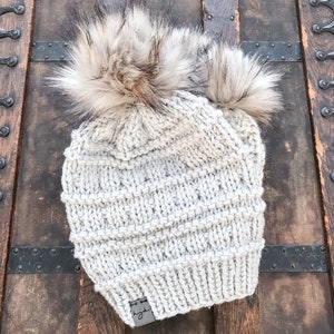 Puede incluir: Un gorro de punto gris con dos pompones esponjosos en la parte superior. El gorro tiene una peque&ntilde;a etiqueta de cuero con la palabra "light" en ella.