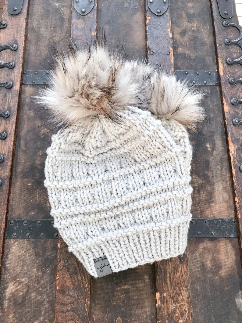 Puede incluir: Un gorro de punto gris con dos pompones esponjosos en la parte superior. El gorro tiene una peque&ntilde;a etiqueta de cuero con la palabra "light" en ella.
