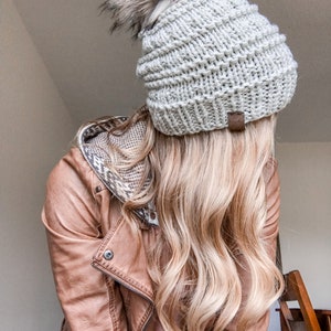 Puede incluir: Una mujer con una chaqueta de cuero marr&oacute;n y una boina de punto blanco con un pomp&oacute;n esponjoso. Su cabello es largo y rubio.