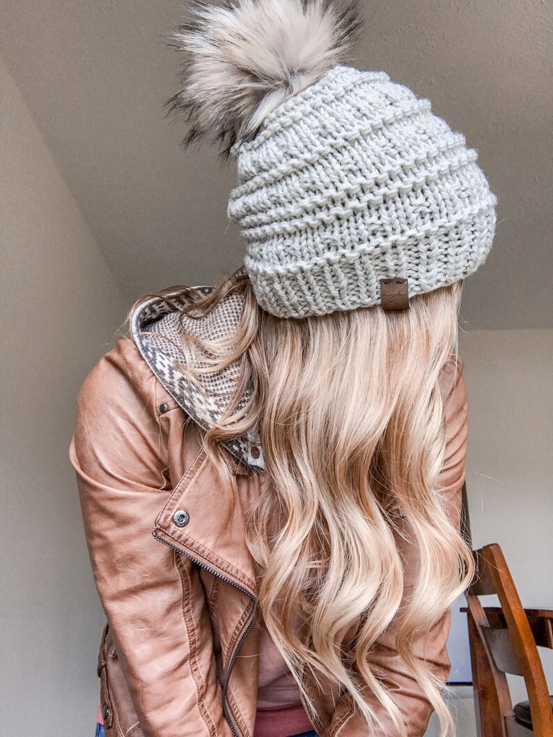 Puede incluir: Una mujer con una chaqueta de cuero marr&oacute;n y una boina de punto blanco con un pomp&oacute;n esponjoso. Su cabello es largo y rubio.