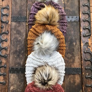 Puede incluir: Cuatro gorros de punto con pompones de piel sint&eacute;tica en varios colores, incluyendo morado, naranja, gris y rojo. Los gorros est&aacute;n apilados uno encima del otro y se muestran sobre un fondo de madera.