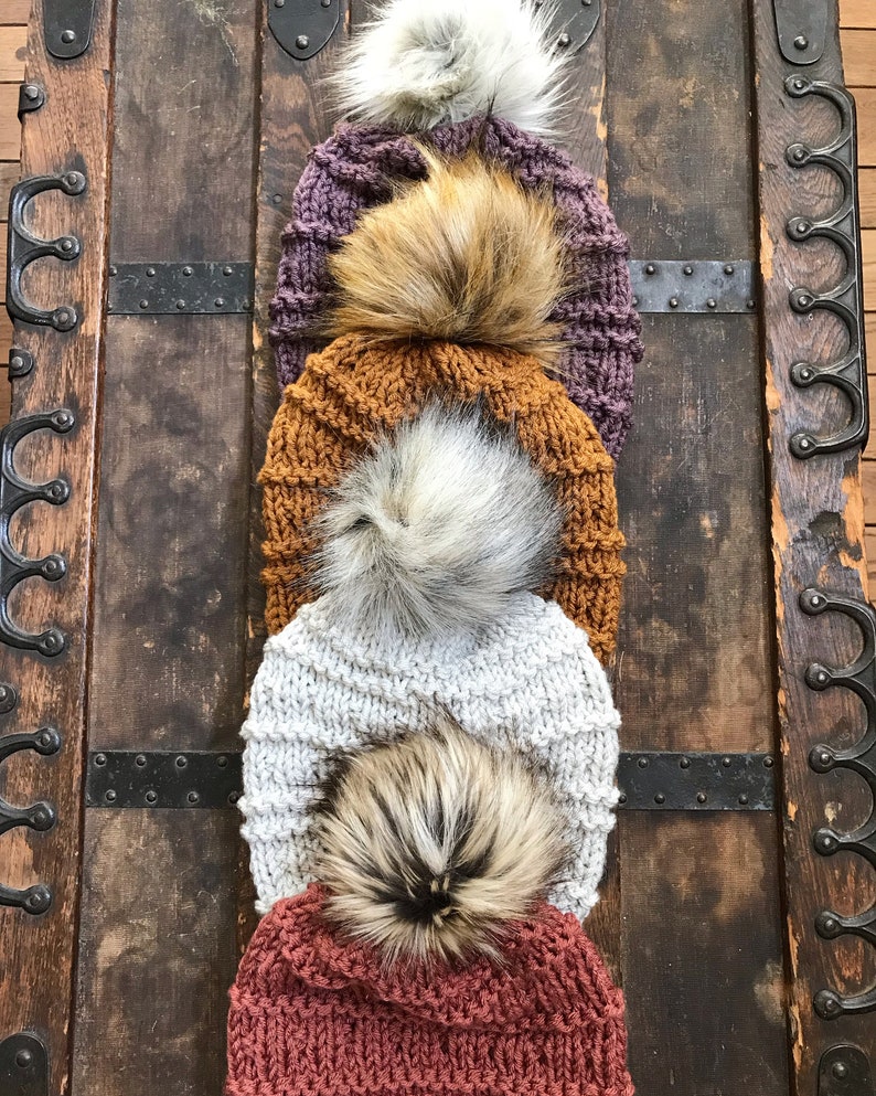 Puede incluir: Cuatro gorros de punto con pompones de piel sint&eacute;tica en varios colores, incluyendo morado, naranja, gris y rojo. Los gorros est&aacute;n apilados uno encima del otro y se muestran sobre un fondo de madera.