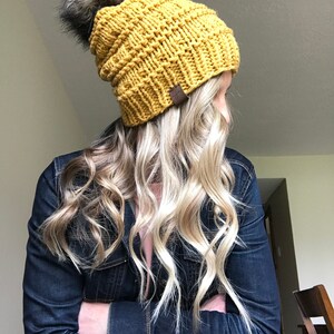 Puede incluir: Un gorro de punto amarillo mostaza con un pomp&oacute;n negro esponjoso. El gorro est&aacute; hecho con un hilo grueso y tiene una textura acanalada.
