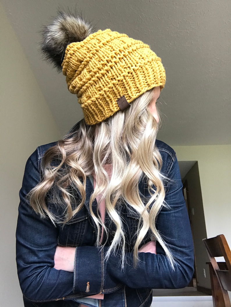 Puede incluir: Un gorro de punto amarillo mostaza con un pomp&oacute;n negro esponjoso. El gorro est&aacute; hecho con un hilo grueso y tiene una textura acanalada.
