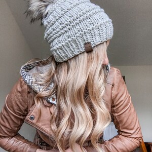 Puede incluir: Un gorro de punto gris con un pomp&oacute;n blanco esponjoso. El gorro tiene una etiqueta de cuero marr&oacute;n con la palabra "Cozy" en ella. El gorro lo lleva una persona con el pelo largo rubio y una chaqueta de cuero marr&oacute;n.