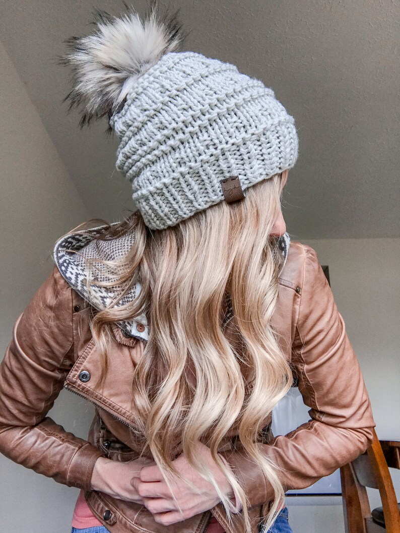Puede incluir: Un gorro de punto gris con un pomp&oacute;n blanco esponjoso. El gorro tiene una etiqueta de cuero marr&oacute;n con la palabra "Cozy" en ella. El gorro lo lleva una persona con el pelo largo rubio y una chaqueta de cuero marr&oacute;n.
