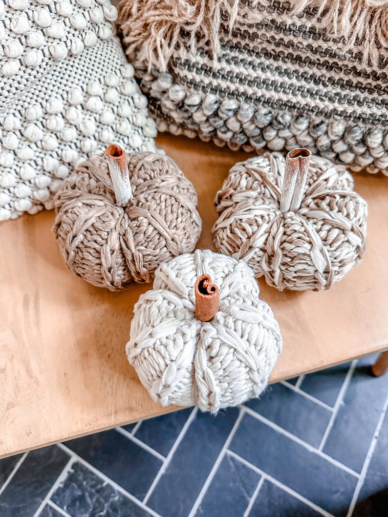 May include: Three knitted pumpkins with cinnamon stick stems. The pumpkins are beige and white and are sitting on a wooden surface.