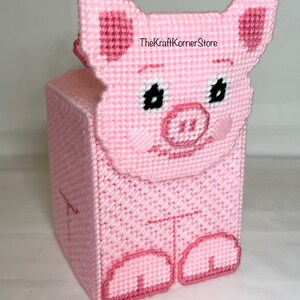 May include: Pink crocheted tissue box cover shaped like a pig. The pig has black eyes, a pink snout, and pink feet.